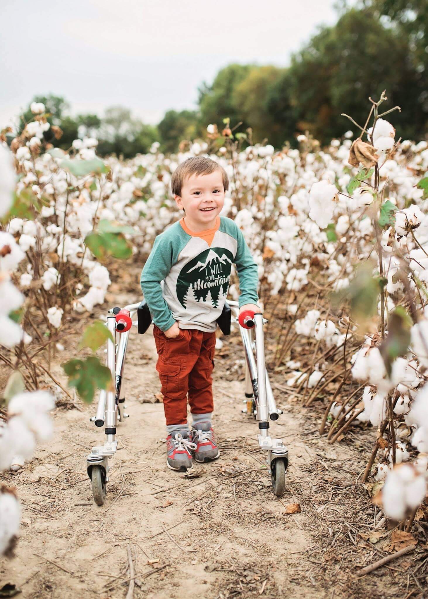 A little boy standing with his walker before he had SDR. He is wearing a blue and white shirt that says: I will move mountains. He is smiling.