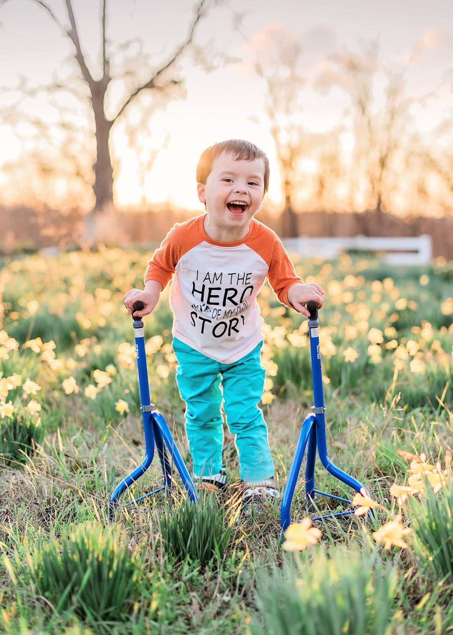 A young boy who had SDR. He is smiling and using blue tripod canes. His shirt says, I am the hero of my own story.
